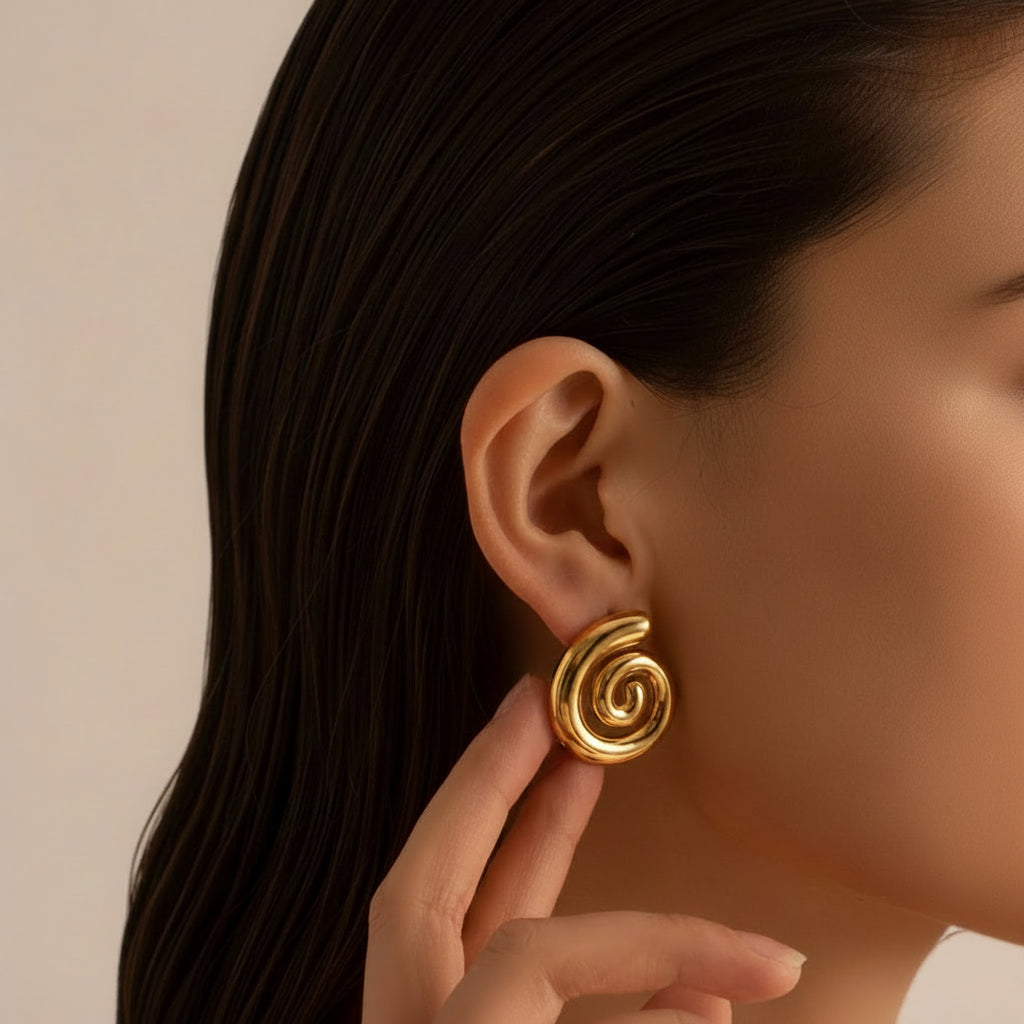 Close-up van een elegante vrouw met donker glanzend haar, die een grote statement gouden spiraal oorbel van CECELYN draagt. Haar hand raakt de oorbel zachtjes aan, waardoor het sculpturale en glanzende design perfect in beeld komt. De warme gouden kleur contrasteert prachtig met haar huid en donkere haren. Een stijlvolle en minimalistische productfoto die de moderne en luxe uitstraling van de spiraal oorbellen uit de nieuwe collectie benadrukt.