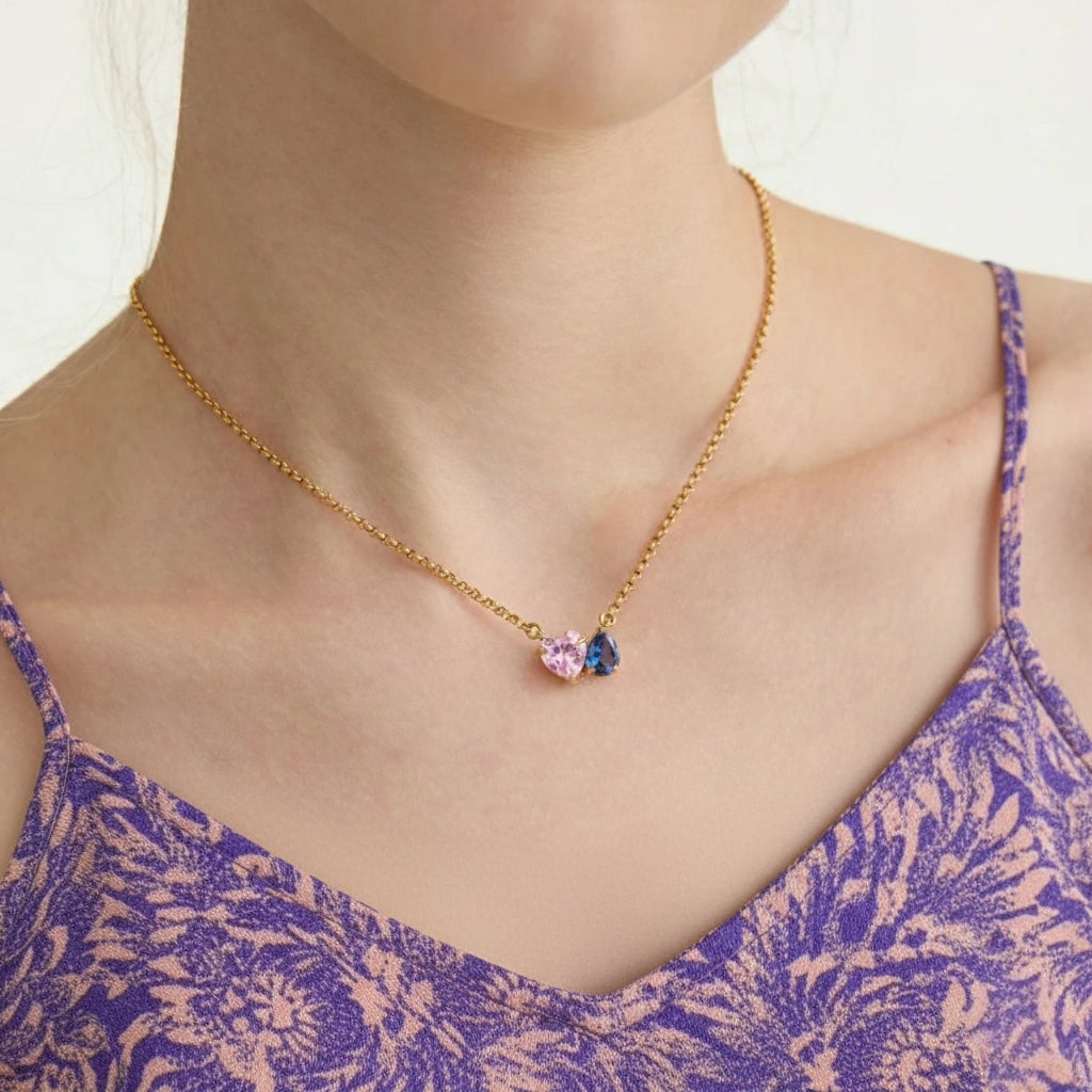 Model draagt minimalistische gouden ketting met roze hart en blauwe steen.