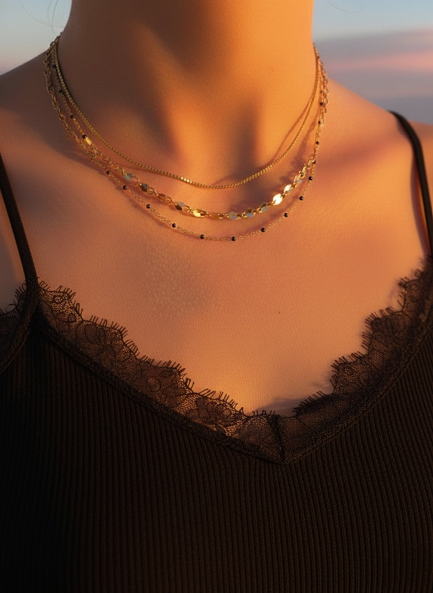 Model draagt een verfijnde goudkleurige gelaagde ketting met zwarte kraaltjes, gecombineerd met een zwarte top met kanten rand in warm zonlicht.