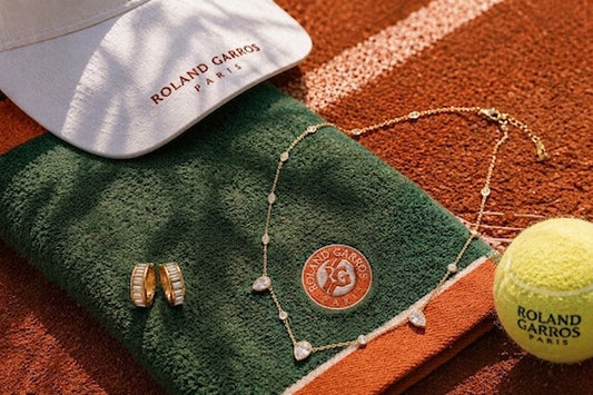 Elegante gouden zirkonia ketting en oorbellen van CECELYN liggend op een officiële Roland Garros handdoek, geflankeerd door een witte zonneklep en een tennisbal op een tennisbaan van gravel.