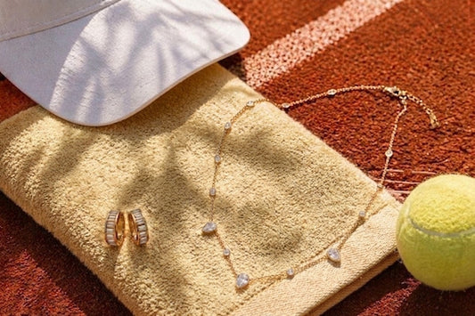 Gouden halsketting met kristallen hangers en bijpassende gouden klapoorringen van CECELYN op een tennisbaan setting.