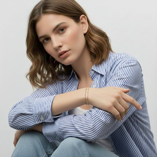 Model draagt een subtiele gouden dubbele schakelarmband met kleine strass steentjes en een blauw gestreepte blouse van CECELYN.