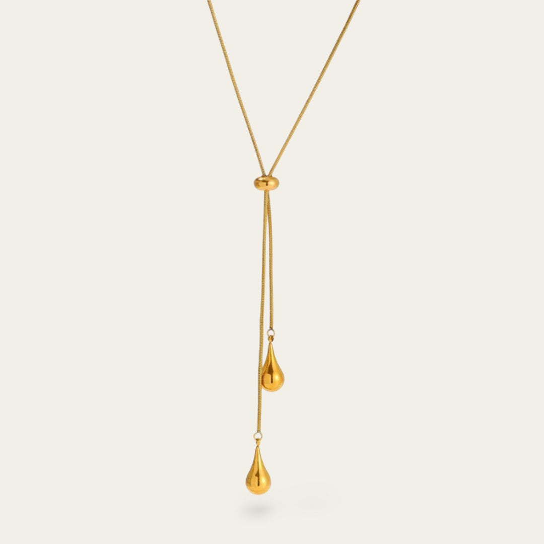 Een close-up van een goudkleurige lariat-ketting tegen een lichte achtergrond. De ketting heeft een fijne slangenschakel en een verstelbare ronde schuifkraal. Aan de uiteinden hangen twee gepolijste, goudkleurige hangers in de vorm van een druppel op verschillende hoogtes.