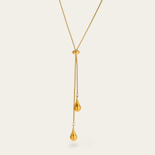 Een close-up van een goudkleurige lariat-ketting tegen een lichte achtergrond. De ketting heeft een fijne slangenschakel en een verstelbare ronde schuifkraal. Aan de uiteinden hangen twee gepolijste, goudkleurige hangers in de vorm van een druppel op verschillende hoogtes.