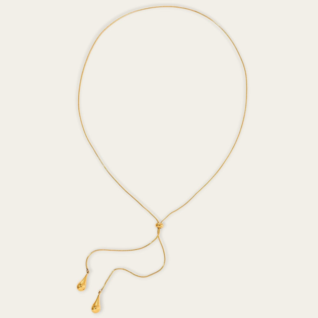 Een bovenaanzicht van de goudkleurige druppelketting, liggend in een cirkelvorm op een neutrale achtergrond. De afbeelding toont duidelijk de volledige lengte van de fijne schakelketting en hoe de twee druppelvormige bedels via de centrale schuifknoop naar beneden vallen.