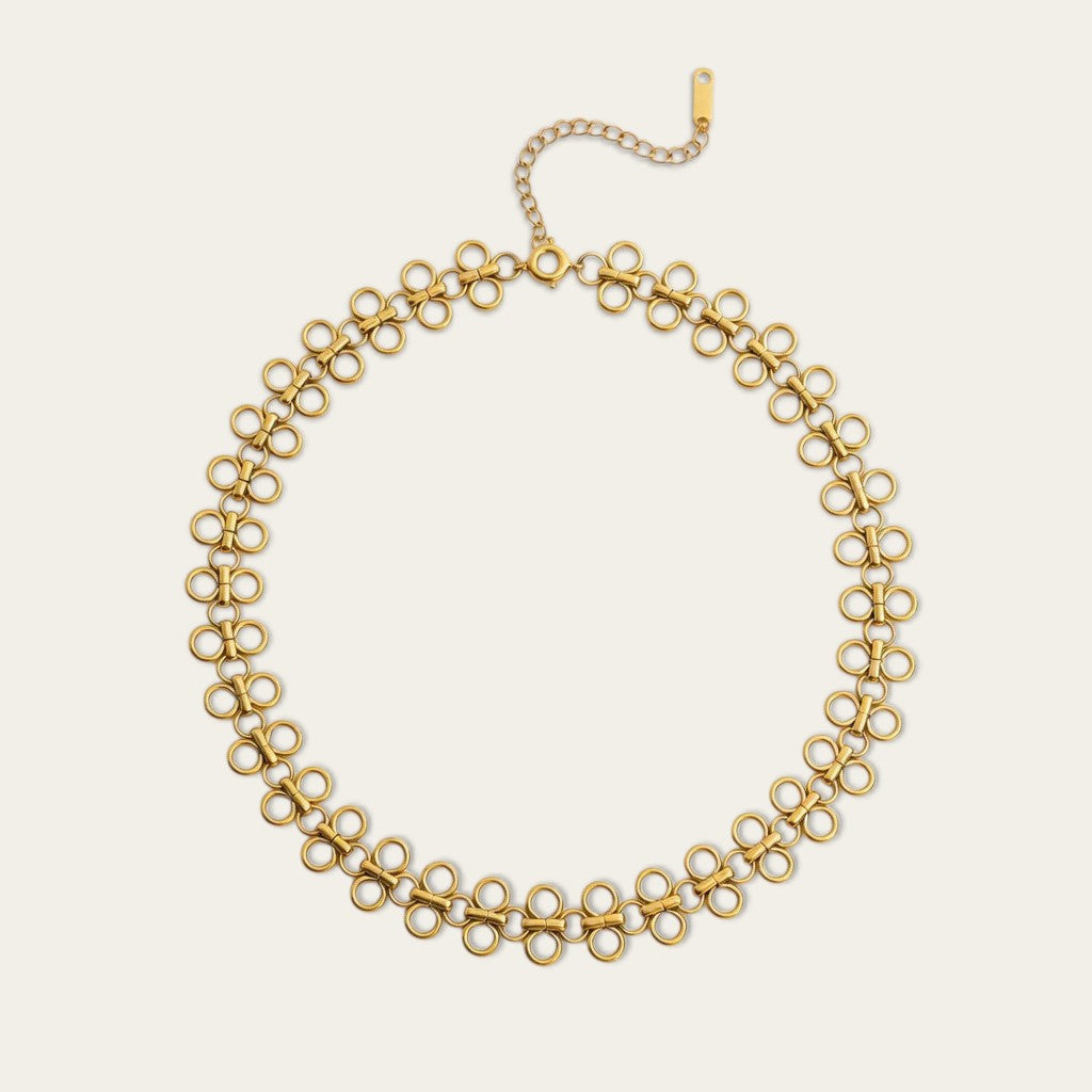 Een goudkleurige filigraan choker ketting plat neergelegd tegen een beige achtergrond. De ketting bestaat uit een herhalend patroon van ronde, in elkaar gevlochten lussen en heeft een verstelbare sluiting met een klein merkplaatje.