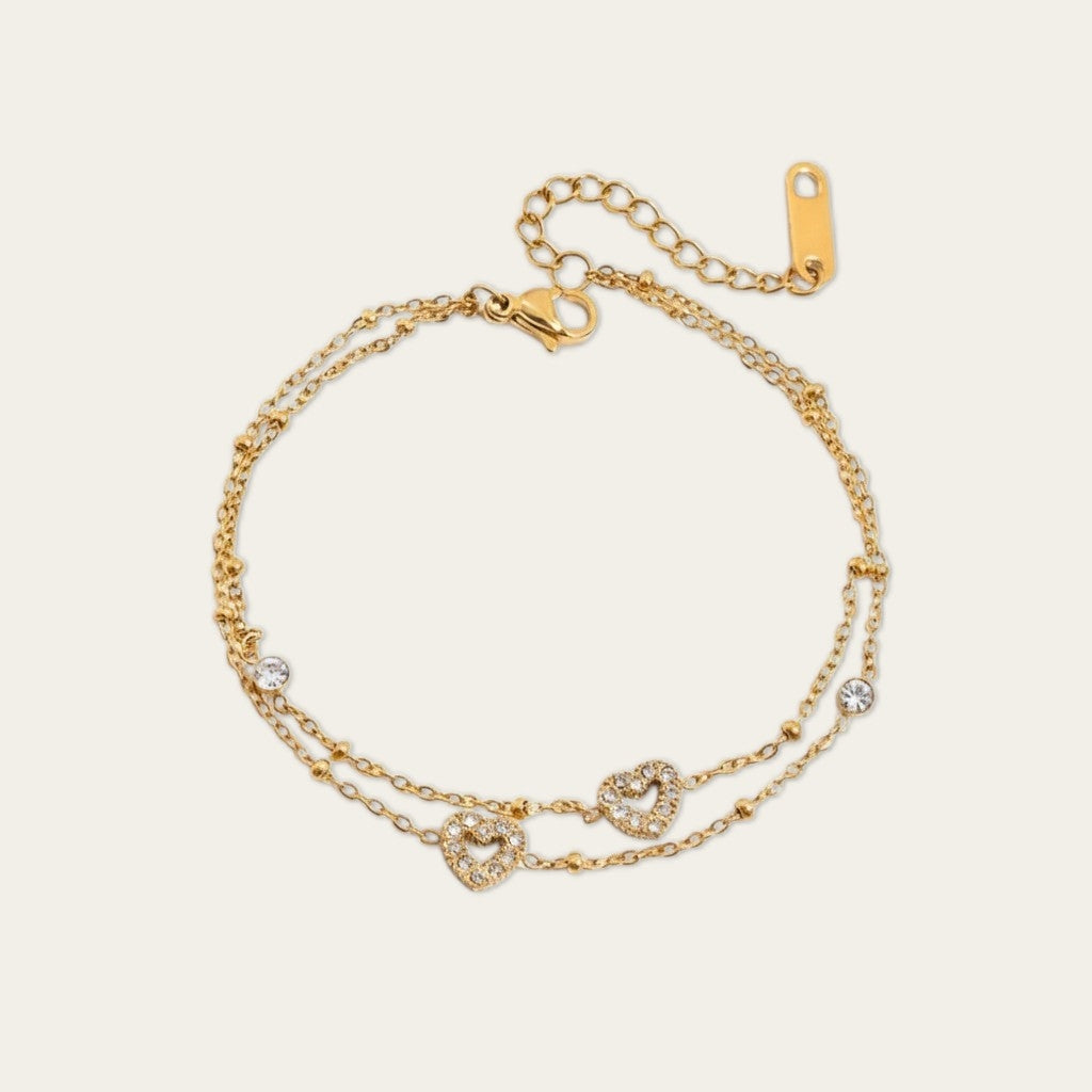Een minimalistische, goudkleurige dubbele armband van CECELYN tegen een egale witte achtergrond. De armband bestaat uit twee fijne, gelaagde kettingen versierd met subtiele goudkleurige bolletjes. Aan de kettingen hangen twee verfijnde, opengewerkte hartjes die volledig zijn ingezet met glinsterende zirkonia steentjes, geflankeerd door twee kleine, ronde strassteentjes. De armband is voorzien van een goudkleurige karabijnsluiting, een verlengketting en een langwerpig merktag.