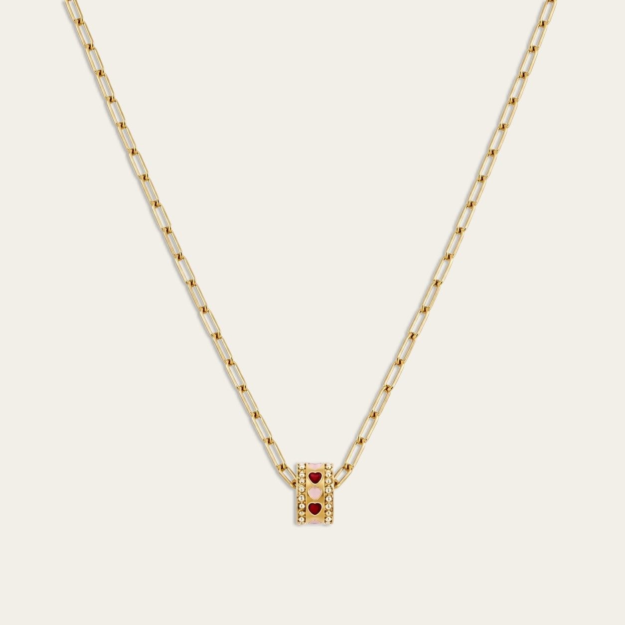 Een close-up studio-opname van een gouden CECELYN paperclip ketting met een cilindervormige bedel tegen een strakke witte achtergrond. De hanger is gedetailleerd afgewerkt met sprankelende steentjes en kleine, ingelegde rode en roze hartjes.