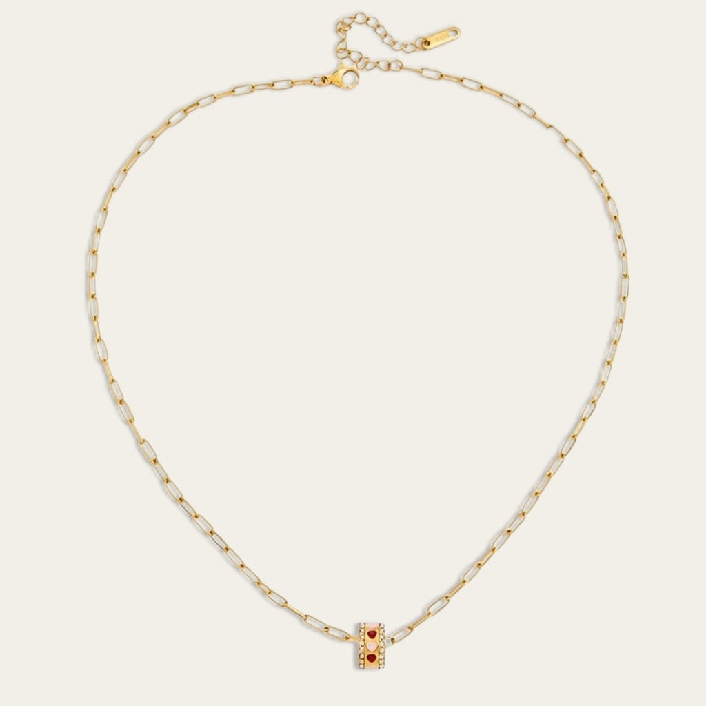 Een minimalistische gouden paperclip ketting van CECELYN met een cilindervormige hanger, plat gefotografeerd tegen een witte achtergrond. De kleine cilinder is versierd met een rij ingezette steentjes en roze en rode hartjes. De ketting heeft een fijne karabijnsluiting met een verlengstukje.