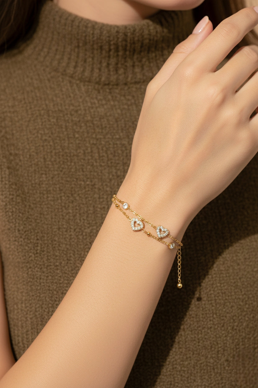 Sfeerbeeld van een model dat de minimalistische, goudkleurige dubbele armband van CECELYN om de pols draagt. De fijngevoelige dubbele ketting met kleine bolletjes, twee opengewerkte zirkonia hartjes en subtiele ronde strassteentjes komt mooi uit tegen de huid. Het model draagt een olijfgroene of bruine gebreide coltrui, wat zorgt voor een warme, modieuze en nonchalante uitstraling.