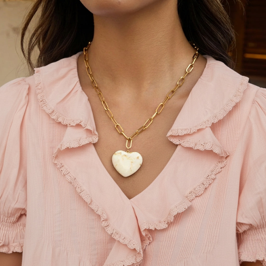 Model draagt minimalistische goudkleurige schakelketting met grote rechthoekige schakels en een hartvormige hanger met een beige marmerpatroon.