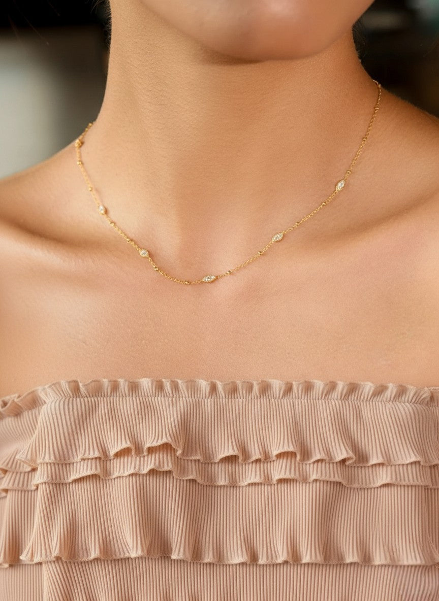 Een close-up opname van het nek- en décolletégedeelte van een vrouwelijk model met een gebruinde huid en donker haar dat naar achteren is gestoken. Ze draagt de goudkleurige station-ketting, die elegant rond haar sleutelbeen valt. De vijf voorste marquise-vormige stations zijn duidelijk zichtbaar en vangen het licht, wat zorgt voor subtiele schittering. Het model draagt een strapless top met plooitjes in een bleekroze/beige kleur.