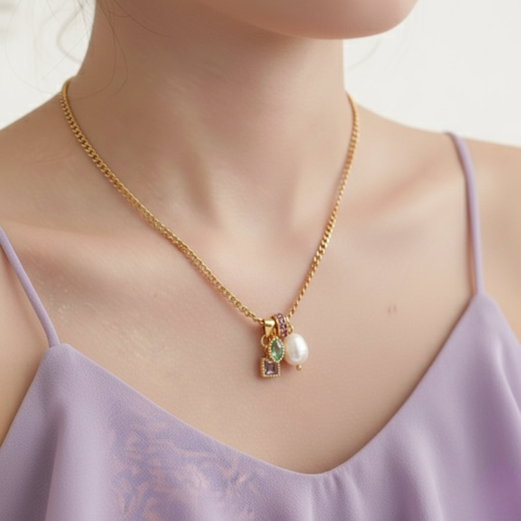 Een foto van de goudkleurige schakelketting gedragen rond de hals van een model. Het model draagt een paarse spaghettitop. De focus ligt op de hals en het decolleté, en op de manier waarop de ketting valt. De vier bedeltjes (zoetwaterparel, paarse steentjes, aqua steen en paarse steen) hangen gecentreerd en rusten net boven de rand van de top. De foto toont de ketting in een realistische context en geeft een indruk van de grootte van de bedeltjes ten opzichte van de drager.