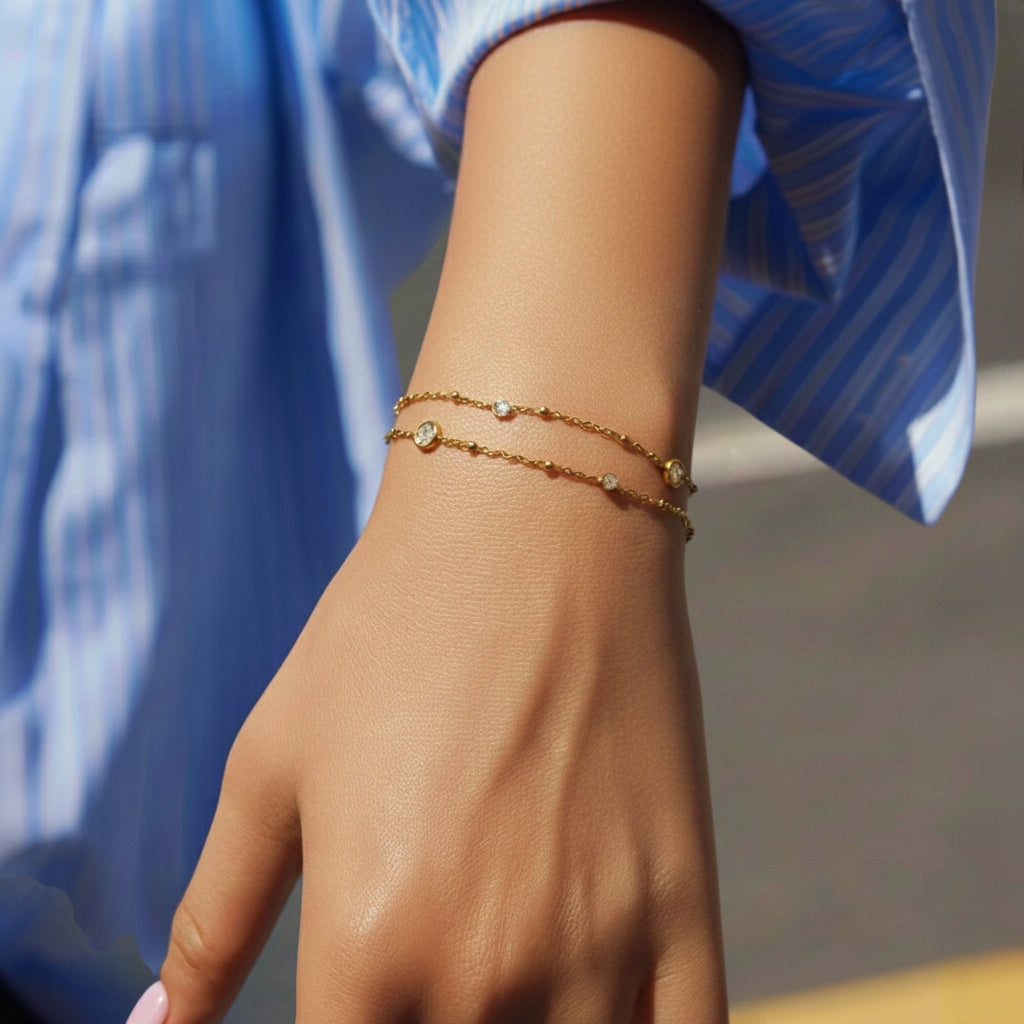 Gedetailleerd: Close-up van de pols van een model dat een minimalistische, goudkleurige dubbele armband van CECELYN draagt. De armband bestaat uit twee zeer fijne kettingen: een klassieke schakelketting en een subtiele bolletjesketting. Beide kettingen zijn bezet met kleine, ronde zettingen met fonkelende strass-steentjes. Het model draagt een blauw met wit gestreept katoenen overhemd, waarvan de mouw nonchalant is opgestroopt.
