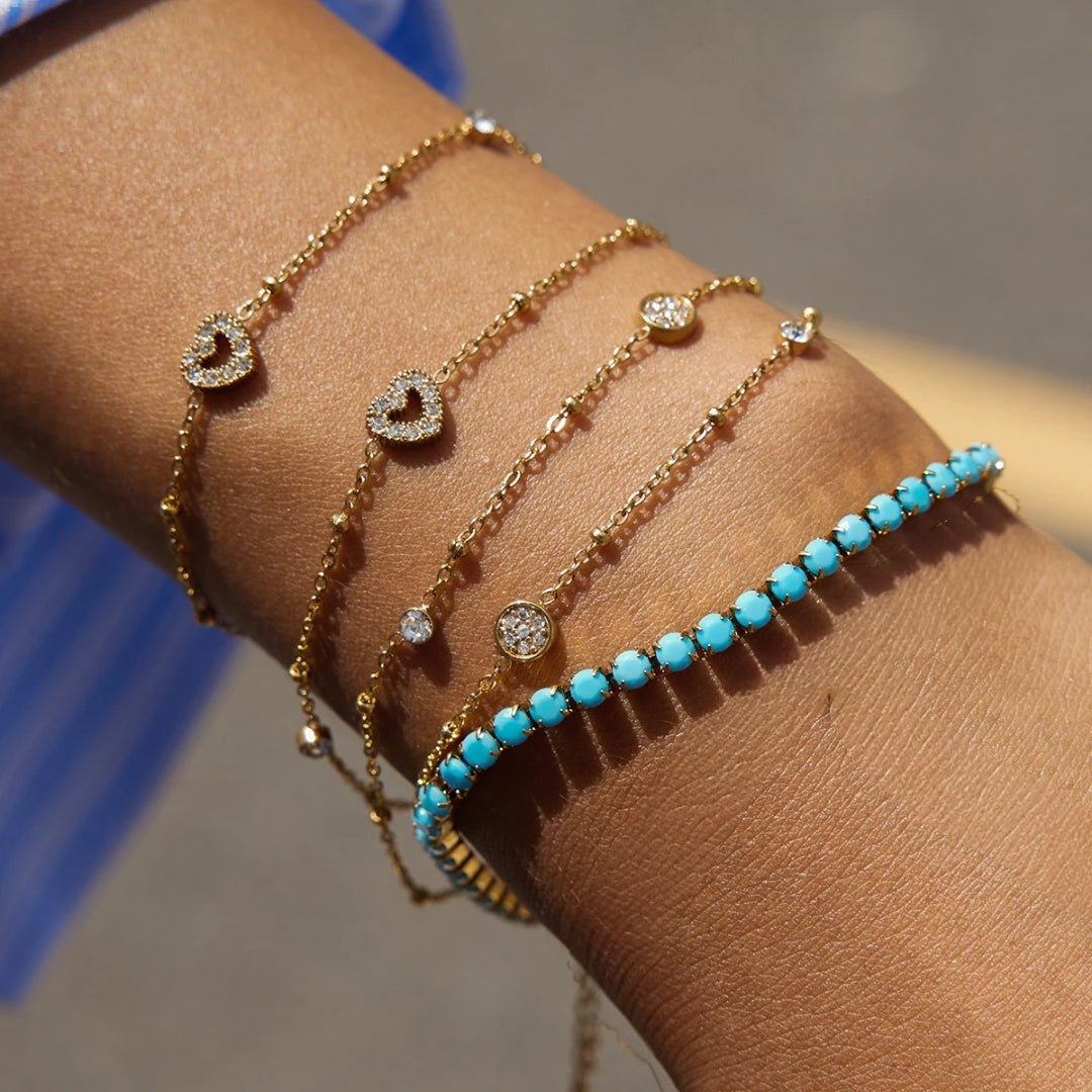 Een gedetailleerde close-up van een 'arm party' aan de pols van een model. Er worden vier verschillende goudkleurige armbanden van CECELYN gelaagd gedragen. Van links naar rechts: een fijne schakelketting met een open hart-bedeltje bezet met strass-steentjes; een fijne bolletjesketting met een identiek hart-bedeltje; een fijne bolletjesketting met drie ronde, ingelegde strass-bedeltjes; en een opvallende turquoise tennisarmband met een rij facetgeslepen stenen in een goudkleurige zetting.