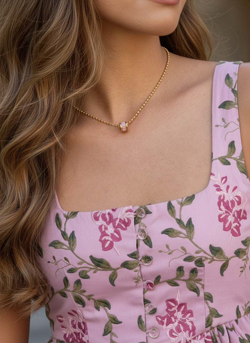 Sfeerbeeld van een model met golvend blond haar, die de goudkleurige cilinder-charm bolletjesketting van Cecelyn draagt. Ze draagt een bijpassende roze bloemenjurk met groene details.