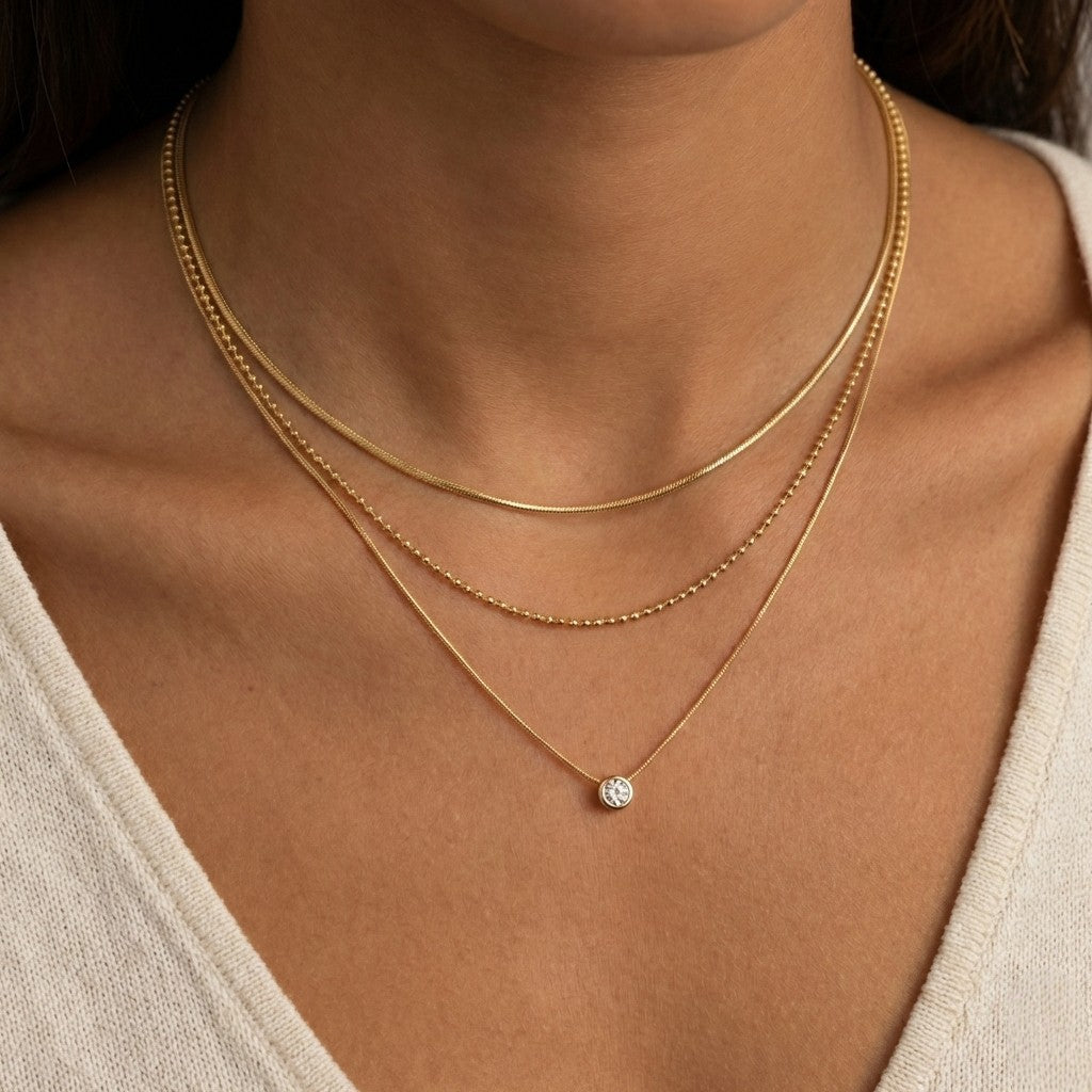 Model draagt een goudkleurige layered necklace set bestaande uit een platte slangenketting, een kralenketting en een fijne schakelketting met een glanzend steentje.