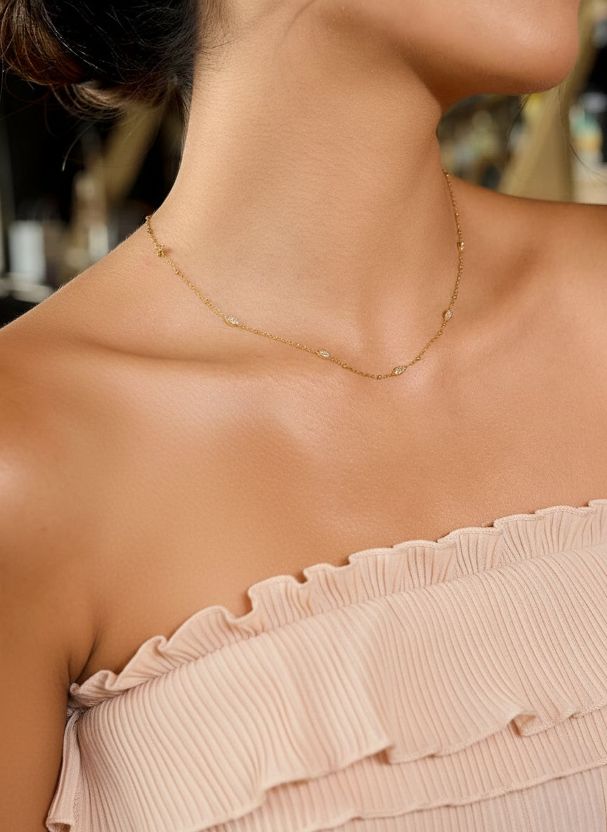 Een close-up opname van het nek- en schoudergedeelte van het vrouwelijke model, genomen vanuit een lichte zijhoek. Deze hoek accentueert de delicate structuur van de goudkleurige ketting terwijl deze over haar sleutelbeen en naar haar schouder loopt. De fijne schittering van de marquise-vormige stations is goed te zien. Het model draagt dezelfde geplooide, strapless top.