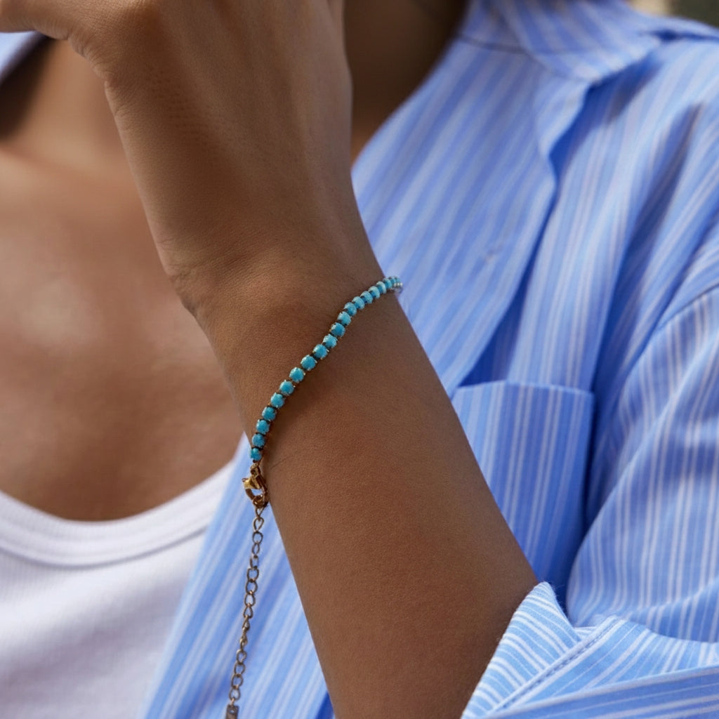 Een close-up lifestyle-foto van de pols van een persoon die de goudkleurige en turquoise tennisarmband van CECELYN draagt. De armband is elegant om de pols gewikkeld, waarbij de opeenvolging van kleine goudkleurige chaton-zettingen en levendig turquoise blauwe steentjes duidelijk zichtbaar is tegen de huid. 