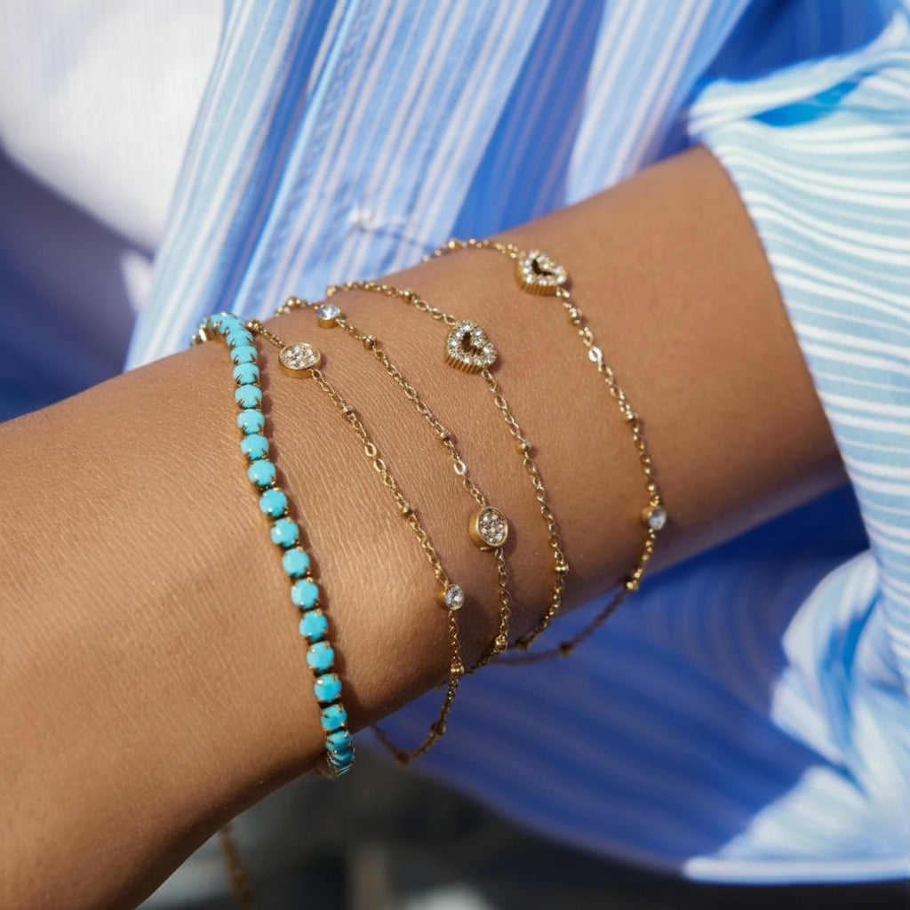 ​Een close-up lifestyle-foto van een gestapelde 'arm candy' sieraadset van CECELYN op de pols van een persoon. Vijf verschillende goudkleurige armbanden zijn elegant gelaagd tegen de achtergrond van een blauw en wit pinstripe overhemd.