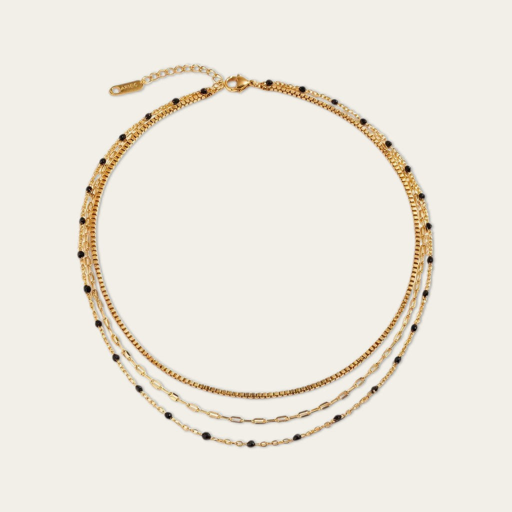 Goudkleurige gelaagde ketting met drie fijne schakels en kleine zwarte kraaltjes, weergegeven op een lichte achtergrond.