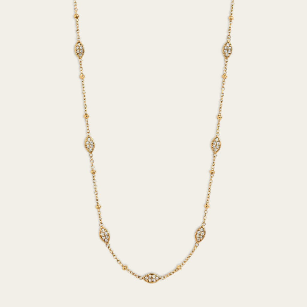 Een Close-up opname van een minimalistische goudkleurige station-ketting tegen een effen, lichte crème achtergrond. De fijne schakelketting is verfraaid met vijf afzonderlijk geplaatste, vaste marquise-vormige stations, elk ingelegd met meerdere kleine, heldere steentjes. De stations zijn gelijkmatig verdeeld over de lengte van de ketting.