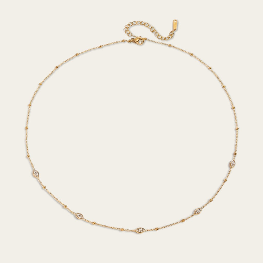 Een lay-flat productfoto van de goudkleurige station-ketting, gerangschikt in een perfecte cirkel tegen een effen, lichte crème achtergrond. Deze afbeelding toont duidelijk de volledige lengte van de ketting, inclusief de karabijnsluiting en de verstelbare verlengketting bovenaan. De vijf marquise-vormige stations ingelegd met steentjes zijn zichtbaar langs de omtrek.