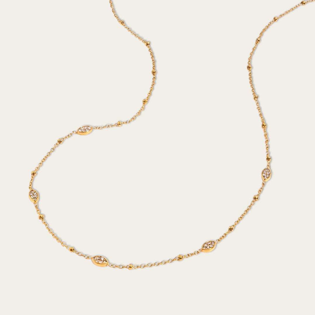Een artistieke close-up productfoto van een gedeelte van de goudkleurige station-ketting, die in een zachte, vloeiende S-vorm is getwist tegen de effen, lichte crème achtergrond. Deze opname legt de fijnere details van de individuele schakels, de kleine metalen bolletjes tussen de stations en de textuur van de marquise-vormige stations ingelegd met zirkonia’s vast.
