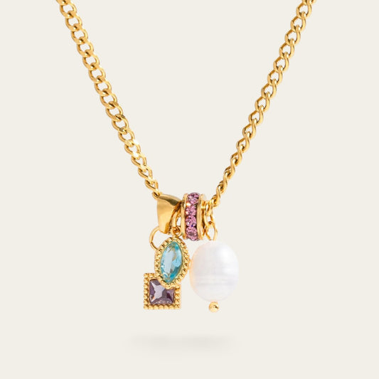 Een gedetailleerde close-up foto die inzoomt op de bedeltjes van de gouden schakelketting. De vier bedeltjes hangen dicht bij elkaar aan een gouden ringetje. Links onderaan zie je een vierkante paarse steen in een bewerkte gouden zetting. Daarboven hangt een marquise-geslepen aqua-blauwe steen in een soortgelijke zetting. Rechts daarvan bevindt zich een rond tussenzetstuk dat volledig is ingelegd met kleine paarse zirkonia steentjes. Geheel rechts hangt een natuurlijk gevormde, witte zoetwaterparel.