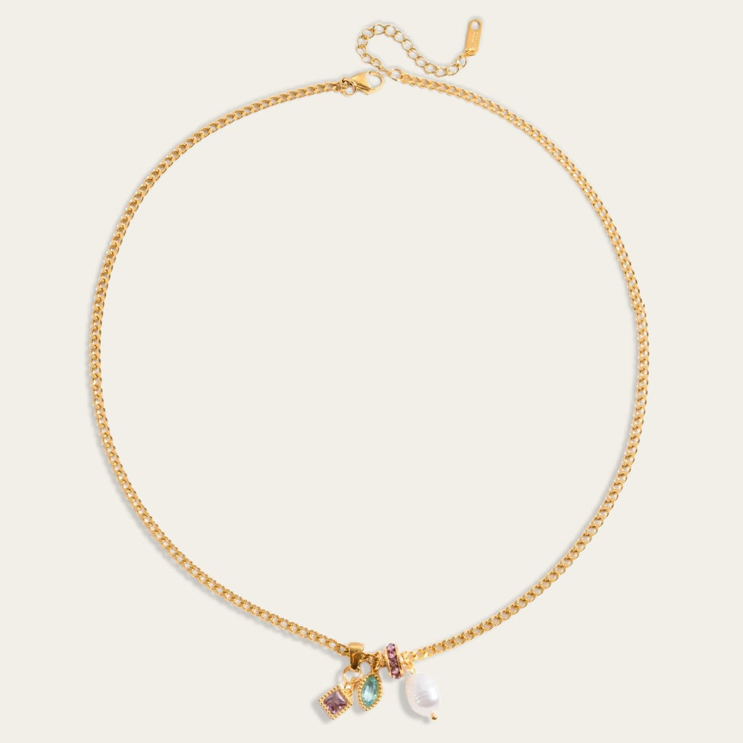 Een plat neergelegde goudkleurige schakelketting op een effen, crèmewitte achtergrond. De ketting vormt een cirkel, met bovenaan de sluiting en het verlengkettinkje. Onderaan, in het midden, hangen de vier verschillende bedeltjes: een grillige witte zoetwaterparel, een rond, met paarse steentjes ingelegd tussenzetstuk, een marquise-geslepen (bladvormige) aqua-blauwe steen en een vierkante paarse steen in een gouden zetting.