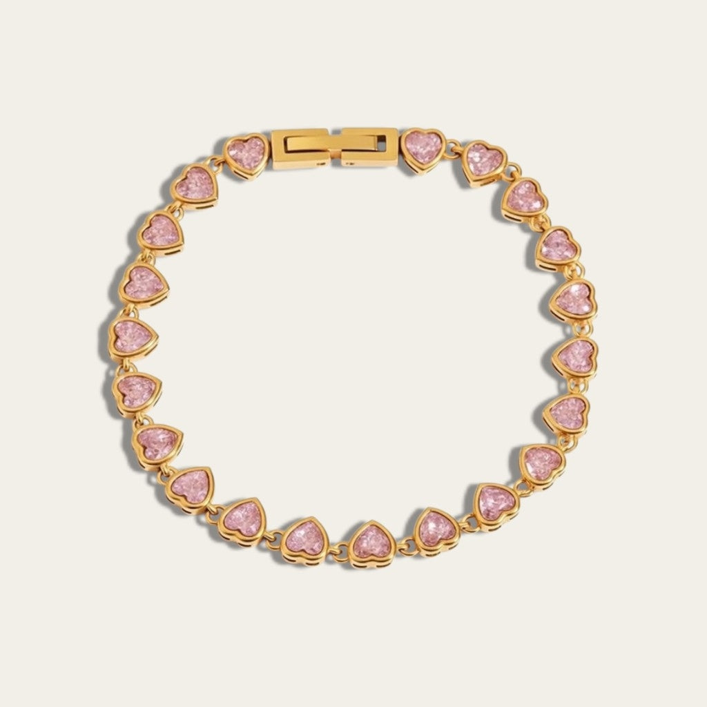 Goudkleurige CECELYN-tennisarmband met lichtroze hartvormige steentjes en een minimalistische kliksluiting op een witte achtergrond.