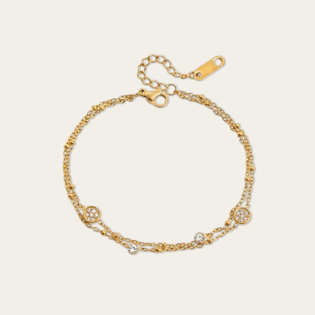 Een verfijnde, goudkleurige dubbele armband van CECELYN tegen een lichte achtergrond. De armband bestaat uit twee fijne, gelaagde kettingen: een subtiele schakelketting en een bolletjesketting. De armband is gedecoreerd met kleine, ronde bedeltjes ingelegd met fonkelende strass-steentjes. Aan het uiteinde zit een goudkleurige karabijnsluiting met een verstelbaar verlengkettingkje en een klein, langwerpig merkplaatje.