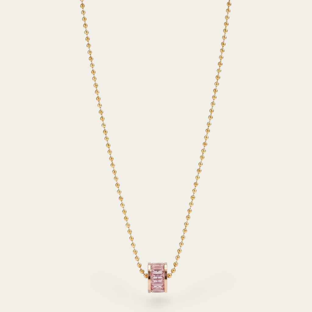 Minimalistische goudkleurige bolletjesketting van CECELYN met een cilindervormige hanger, ingezet met zachtroze kristallen, verticaal gefotografeerd tegen een effen, lichte achtergrond.