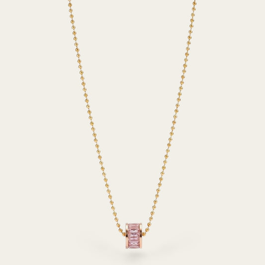 Minimalistische goudkleurige bolletjesketting van CECELYN met een cilindervormige hanger, ingezet met zachtroze kristallen, verticaal gefotografeerd tegen een effen, lichte achtergrond.