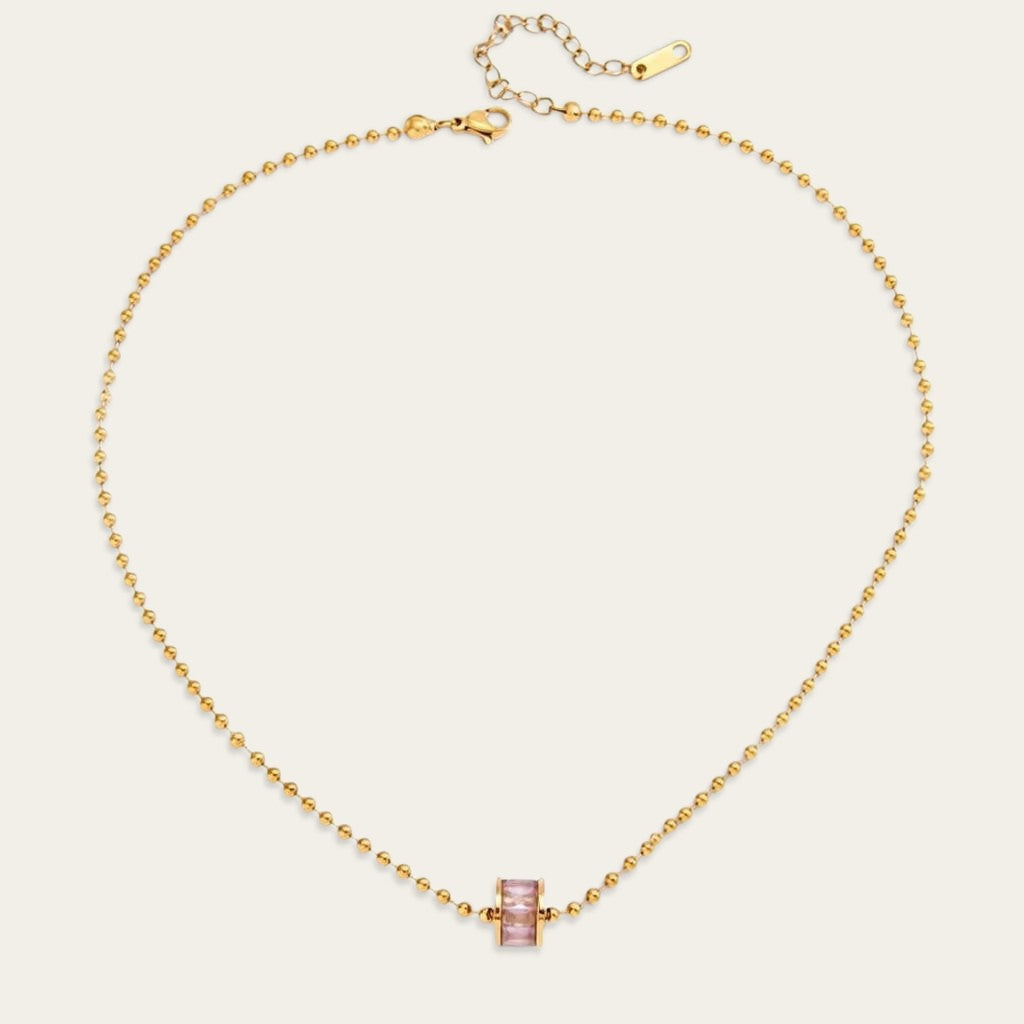 Platte bovenaanzicht van een goudkleurige ballchain ketting, inclusief de karabijnsluiting en verstelketting, met de roze cilindervormige hanger gecentreerd op een witte ondergrond.