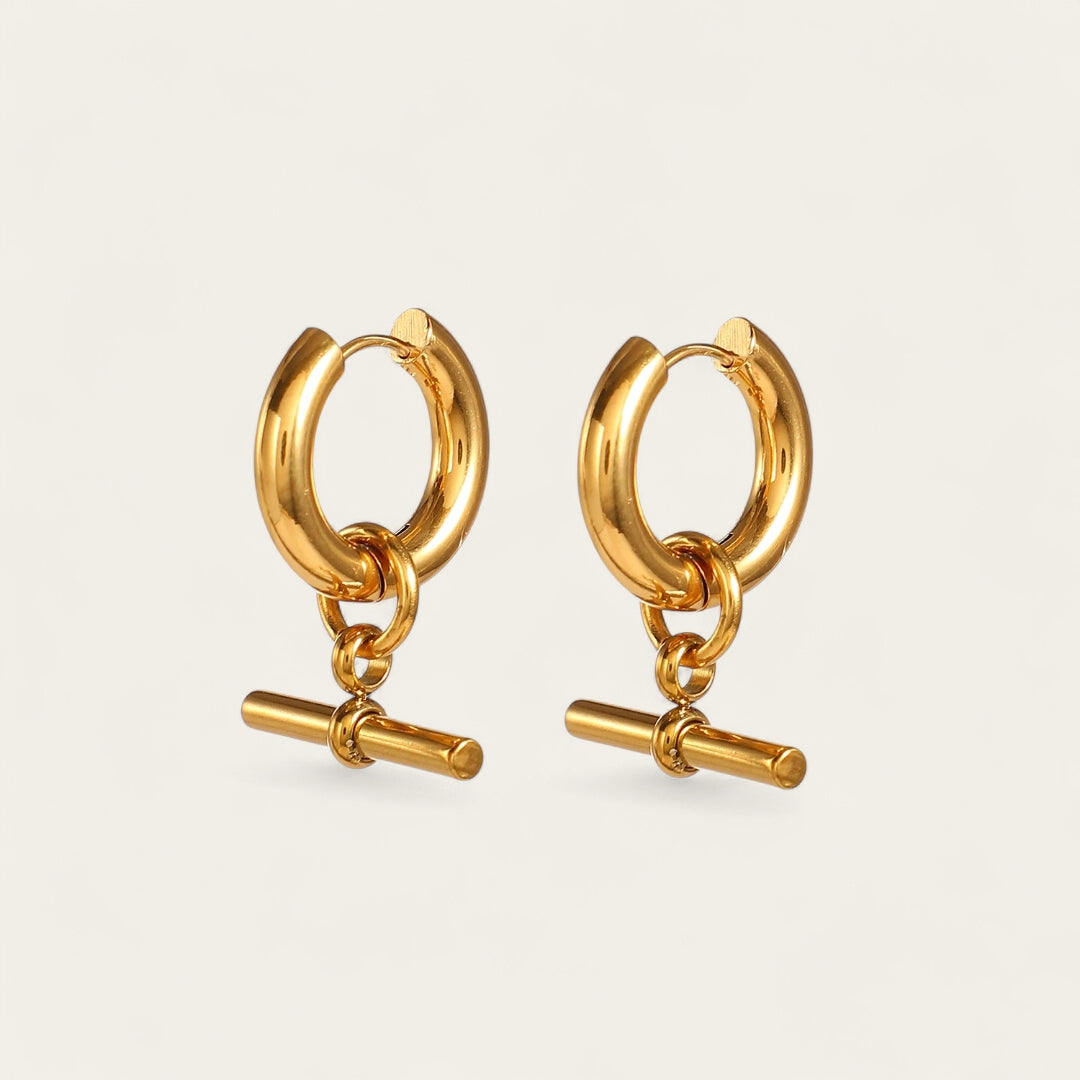 Paar gouden chunky hoop oorbellen met hangende T-bar bedels op een beige achtergrond.