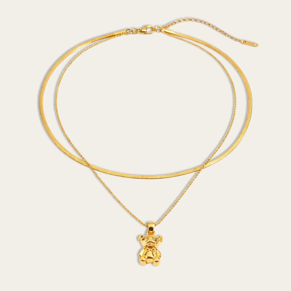 Gelaagde goudkleurige ketting met een gladde snake chain en een fijne bolletjesketting met een glanzende teddybeer hanger.
