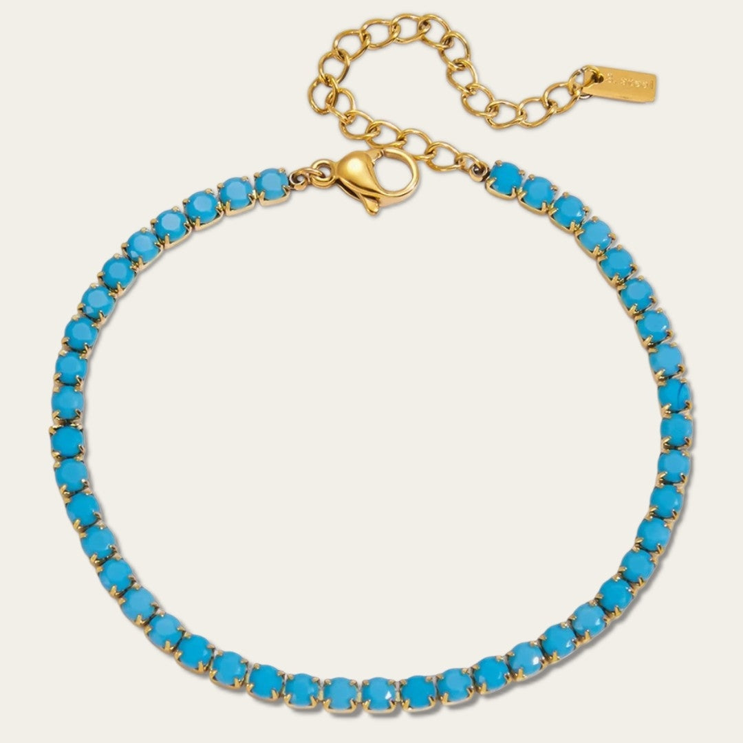 Een gedetailleerde, close-up productfoto van een platliggende goudkleurige tennisarmband van CECELYN op een egale, neutrale lichtgrijze achtergrond. De armband is flexibel en cirkelvormig gerangschikt, gevormd door een aaneenschakeling van tientallen kleine, ronde, goudkleurige chaton-zettingen met pootjes. Elke afzonderlijke zetting bevat een levendig, intens turquoise blauw steentje met een gladde, cabochon-achtige afwerking.